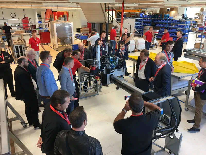 Des visiteurs munis d'un badge entourent une machine de découpe jet d'eau dans l'atelier, pendant qu'un collaborateur en polo rouge donne des explications